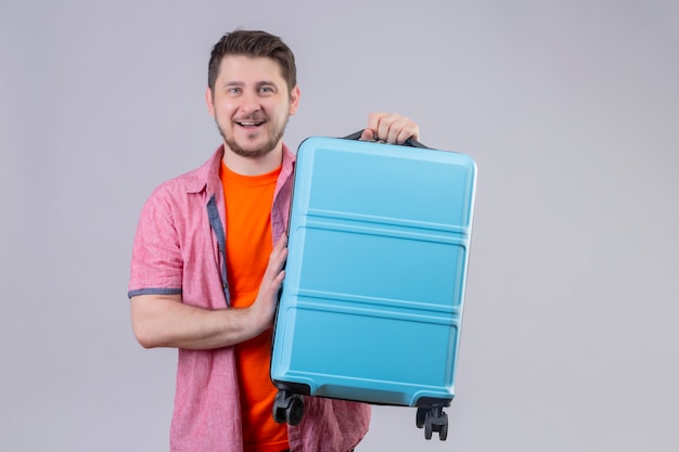 Hombre joven viajero que sostiene la maleta azul mirando a la cámara sonriendo alegremente positivo y feliz de pie sobre fondo blanco.