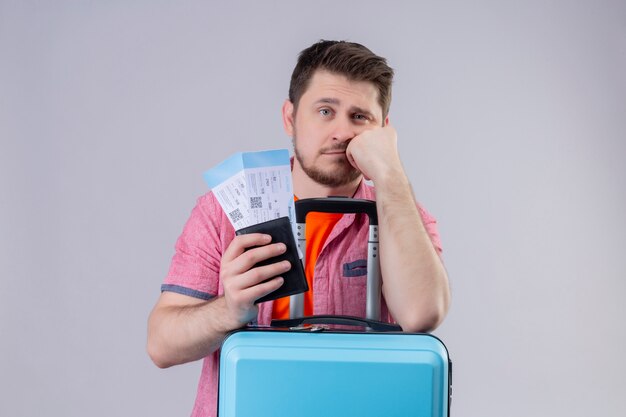 Hombre joven viajero con sombrero de verano sosteniendo billetes de avión y maleta mirando a cámara cansado y aburrido de pie sobre fondo blanco aislado