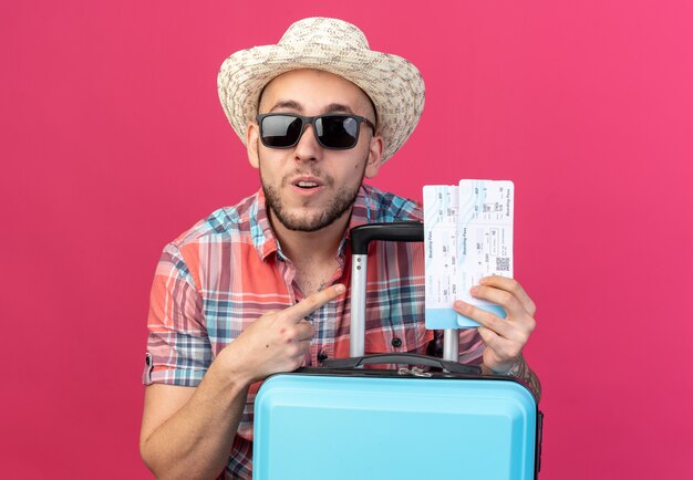 Hombre joven viajero sorprendido con sombrero de playa de paja en gafas de sol sosteniendo y apuntando a los billetes de avión de pie detrás de la maleta aislada en la pared rosa con espacio de copia