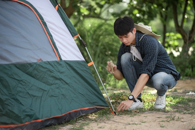 Hombre joven viajero usa una piedra para golpear las clavijas de la tienda en el bosque durante el viaje de campamento en las vacaciones de verano
