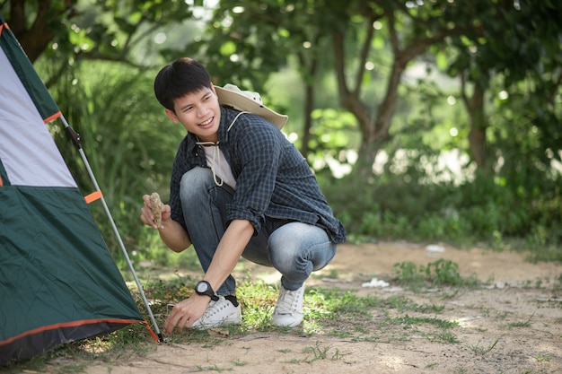 Hombre joven viajero usa una piedra para golpear las clavijas de la tienda en el bosque durante el viaje de campamento en las vacaciones de verano