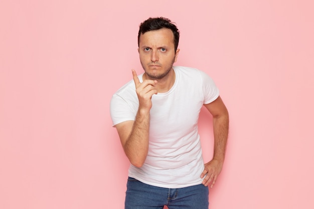 Un hombre joven de vista frontal en camiseta blanca y jeans azul amenazante en el escritorio rosa hombre color emoción pose