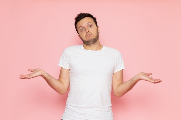 Un hombre joven de vista frontal en camiseta blanca y pantalones vaqueros azules posando con expresión confusa en la pose de emoción de color de hombre de escritorio rosa