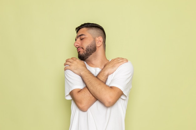 Un hombre joven de vista frontal en camiseta blanca posando y abrazándose a sí mismo