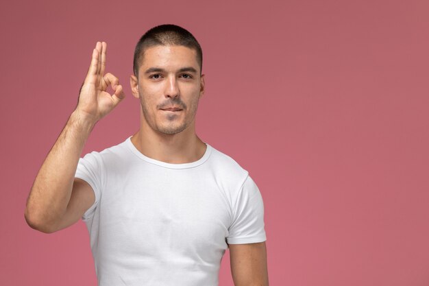 Hombre joven de vista frontal en camiseta blanca posando con la mano levantada sobre el fondo rosa