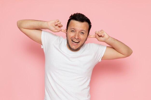 Un hombre joven de vista frontal en camiseta blanca que cubre sus oídos en el escritorio rosa hombre color emoción pose