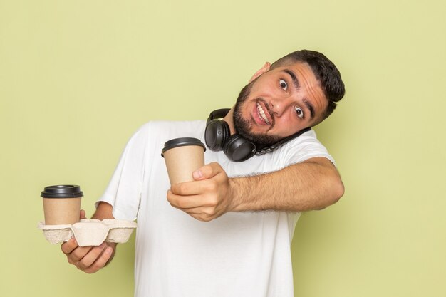 Un hombre joven de vista frontal en camiseta blanca sosteniendo tazas de café y hablando por teléfono