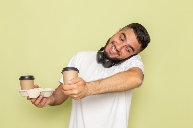 Un hombre joven de vista frontal en camiseta blanca sosteniendo tazas de café mientras habla por teléfono