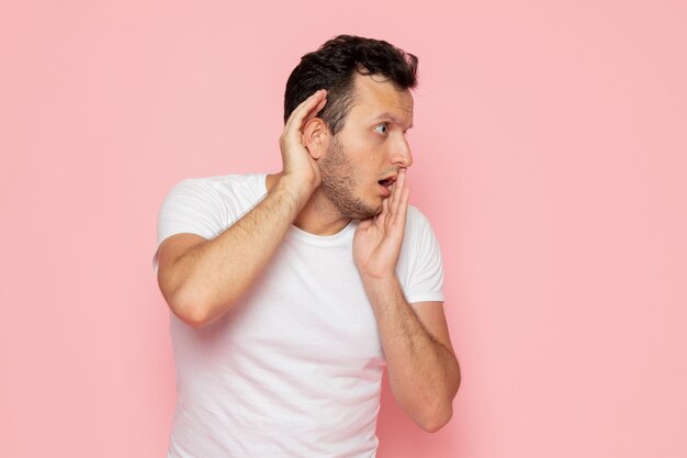 Un hombre joven de vista frontal en camiseta blanca tratando de escuchar en el escritorio rosa hombre color emoción pose