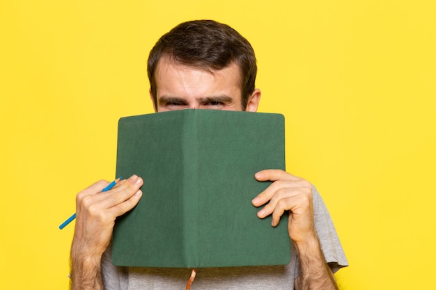 Un hombre joven de vista frontal en camiseta gris con cuaderno verde en la pared amarilla hombre color modelo emoción ropa