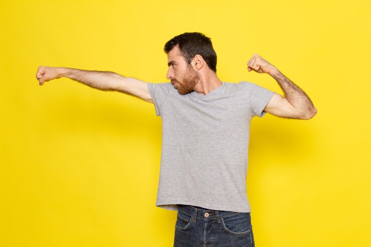 Foto gratuito un hombre joven de vista frontal en camiseta gris lanzando una patada en la pared amarilla hombre expresión emoción modelo de color