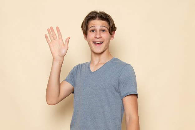 Un hombre joven de vista frontal en camiseta gris posando agitando su mano con una sonrisa