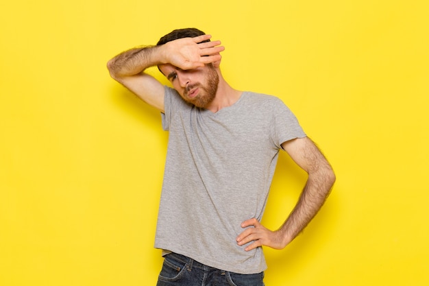 Un hombre joven de vista frontal en camiseta gris posando con expresión cansada en el modelo de color de hombre de pared amarilla