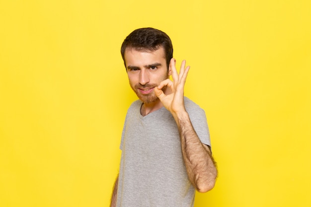Un hombre joven de vista frontal en camiseta gris que muestra bien firmar en la pared amarilla hombre color modelo emoción ropa