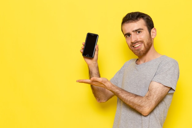 Un hombre joven de vista frontal en camiseta gris con smartphone con sonrisa en el modelo de color de hombre de pared amarilla