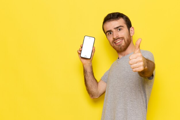 Un hombre joven de vista frontal en camiseta gris con smartphone con sonrisa en el modelo de color de hombre de pared amarilla