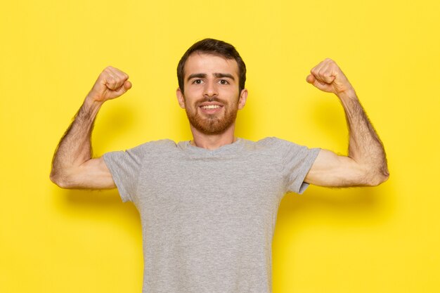 Un hombre joven de vista frontal en camiseta gris sonriendo y flexionando en la pared amarilla hombre color modelo emoción ropa