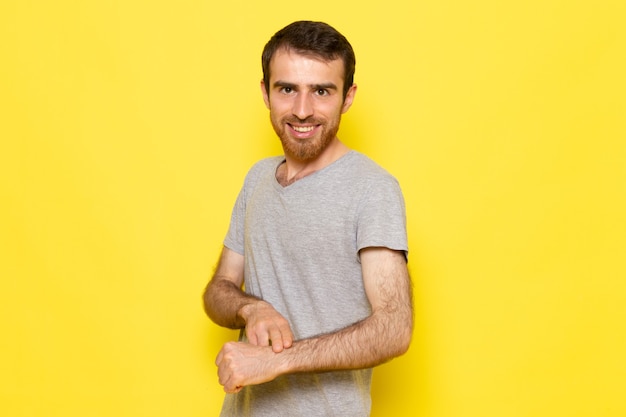 Un hombre joven de vista frontal en camiseta gris sonriendo y señalando en su muñeca en la pared amarilla hombre color modelo emoción ropa