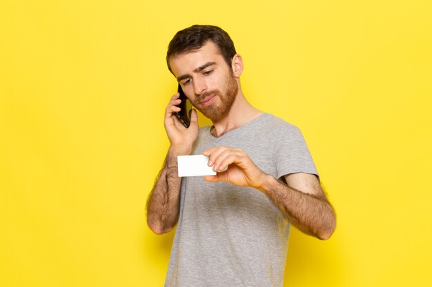 Un hombre joven de vista frontal en camiseta gris sosteniendo una tarjeta blanca y hablando por teléfono en la pared amarilla hombre expresión emoción modelo de color