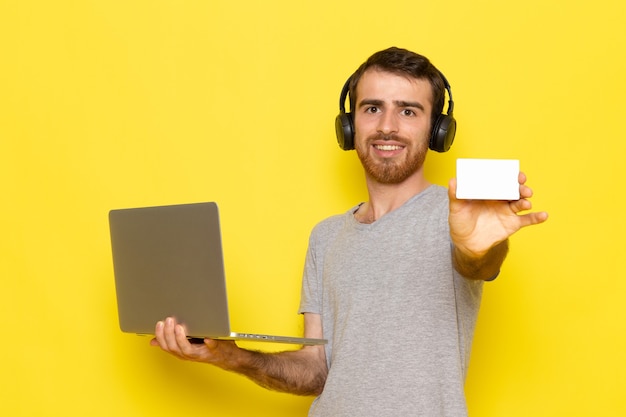 Un hombre joven de vista frontal en camiseta gris sosteniendo una tarjeta blanca y usando una computadora portátil con una sonrisa en la pared amarilla hombre expresión emoción modelo de color