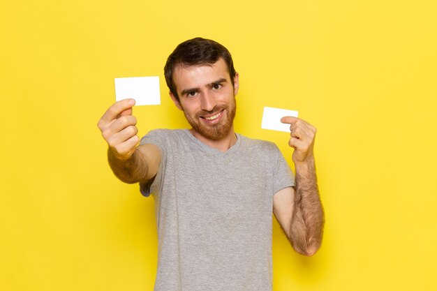 Un hombre joven de vista frontal en camiseta gris sosteniendo tarjetas blancas en la pared amarilla hombre expresión emoción modelo de color
