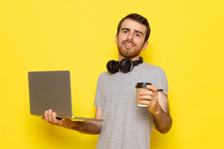 Foto gratuito un hombre joven de vista frontal en camiseta gris sosteniendo una taza de café y usando una computadora portátil con una sonrisa en la pared amarilla hombre expresión emoción modelo de color