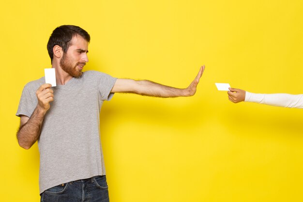 Un hombre joven de vista frontal en camiseta gris con tarjeta blanca rechazando otra tarjeta blanca en la pared amarilla hombre color modelo emoción ropa