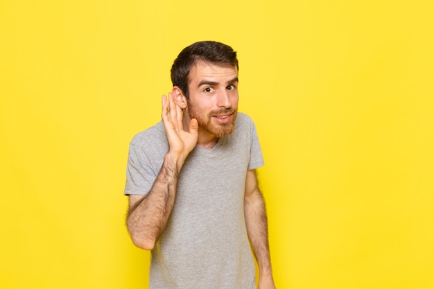 Un hombre joven de vista frontal en camiseta gris tratando de escuchar en la pared amarilla hombre color modelo emoción ropa