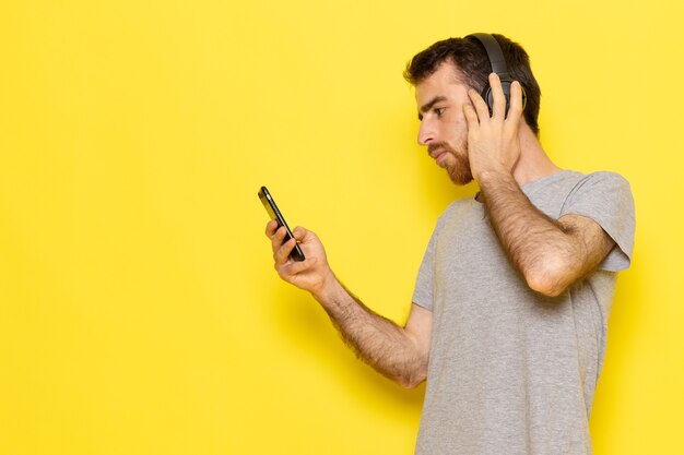 Un hombre joven de vista frontal en camiseta gris usando el teléfono y escuchando música en la pared amarilla hombre expresión emoción modelo de color