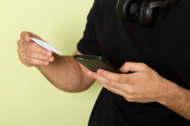 Hombre joven de vista frontal en camiseta negra sosteniendo una tarjeta blanca y usando un teléfono en verde