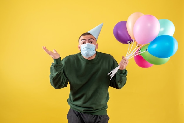 Hombre joven de vista frontal sosteniendo globos de colores en máscara en amarillo