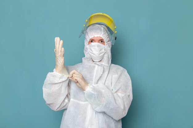 Un hombre joven de vista frontal en traje especial blanco y casco especial amarillo con guantes blancos en la pared azul traje de hombre peligro equipo especial color