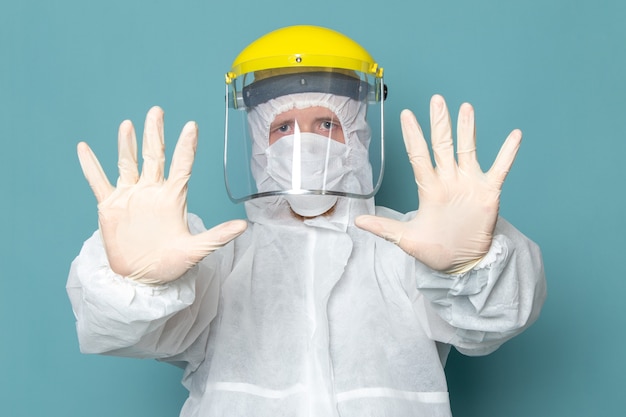 Un hombre joven de vista frontal en traje especial blanco y casco especial amarillo mostrando sus manos en la pared azul traje de hombre peligro equipo especial color