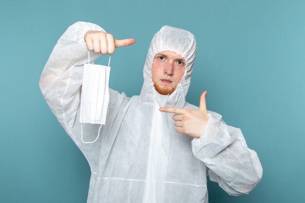 Un hombre joven de vista frontal en traje especial blanco mostrando máscara protectora estéril en la pared azul traje de hombre peligro color de equipo especial