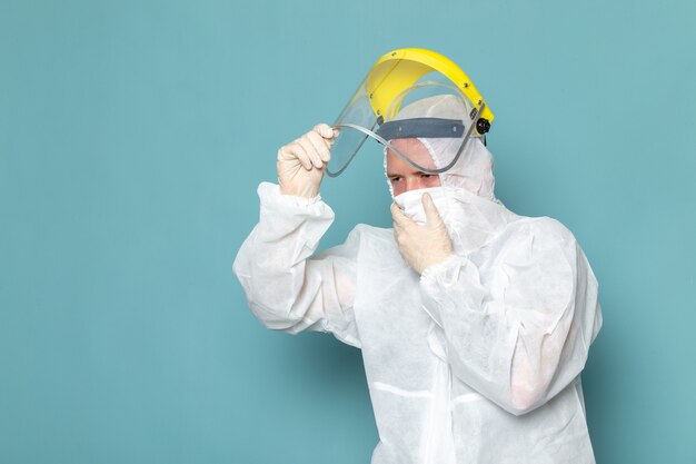 Un hombre joven de vista frontal en traje especial blanco quitándose su casco especial amarillo en la pared azul traje de hombre peligro equipo especial color