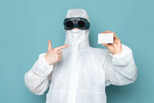 Un hombre joven de vista frontal en traje especial blanco y sosteniendo una tarjeta blanca en la pared azul traje de hombre peligro color de equipo especial