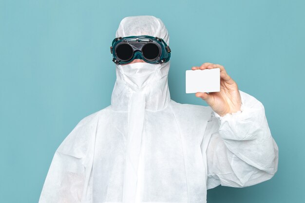 Un hombre joven de vista frontal en traje especial blanco y sosteniendo una tarjeta blanca en la pared azul traje de hombre peligro color de equipo especial