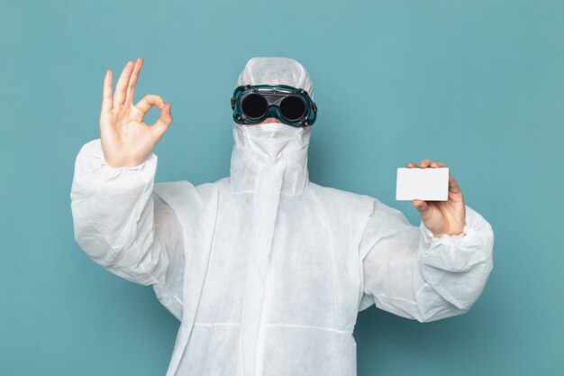 Un hombre joven de vista frontal en traje especial blanco y sosteniendo una tarjeta blanca en la pared azul traje de hombre peligro equipo especial químicos de color
