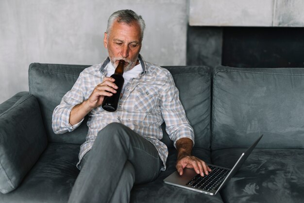 Hombre jubilado bebiendo cerveza de botella usando laptop