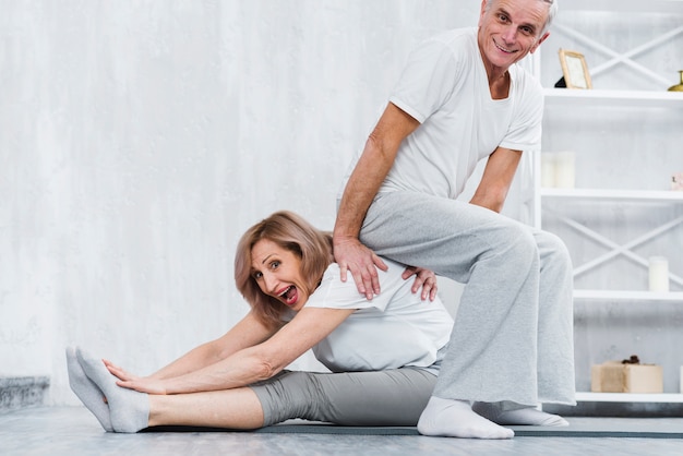 Hombre juguetón sentado en la espalda de su esposa mientras hace yoga en casa