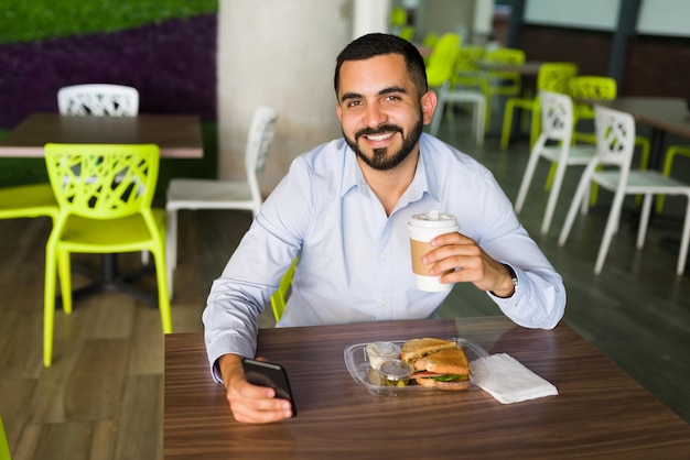 Hombre latino de unos 30 años sonriendo y haciendo contacto visual mientras bebe café y un sándwich durante su almuerzo en el patio de comidas