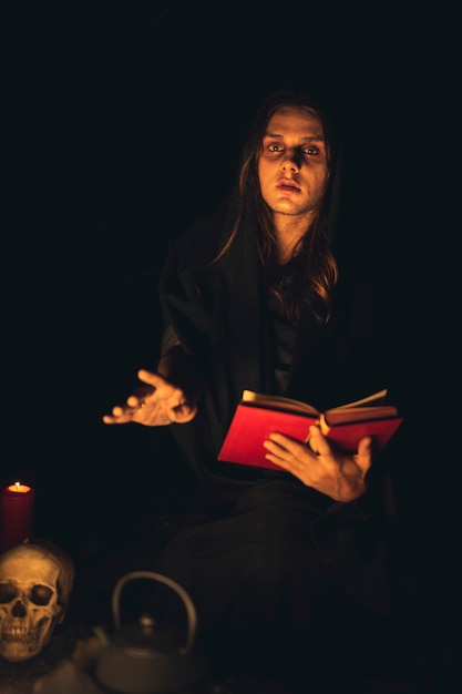 Hombre leyendo un libro de hechizos rojos en la oscuridad y mirando a cámara