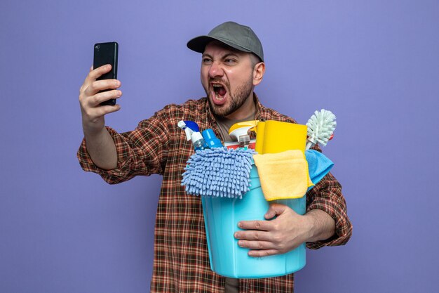 Hombre limpiador eslavo molesto sosteniendo equipo de limpieza y gritando a alguien que mira el teléfono