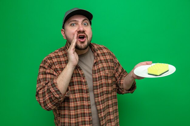 Hombre limpiador eslavo sorprendido manteniendo la mano cerca de la boca y sosteniendo la esponja en el plato