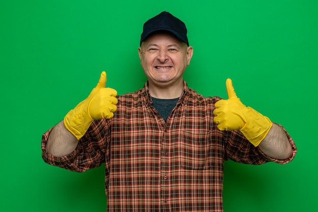 Hombre de limpieza en camisa a cuadros y gorra con guantes de goma mirando feliz y alegre mostrando los pulgares para arriba sonriendo ampliamente