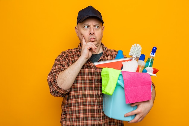 Hombre de limpieza en camisa a cuadros y gorra sosteniendo un balde con herramientas de limpieza mirando a un lado con el ceño fruncido desconcertado de pie sobre fondo naranja