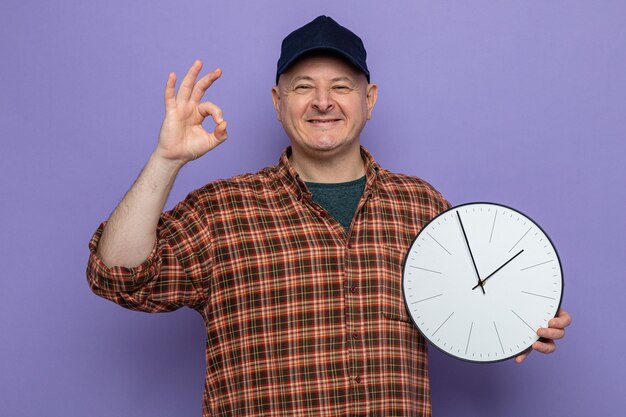 Hombre de limpieza en camisa a cuadros y gorra sosteniendo el reloj mirando a la cámara sonriendo alegremente haciendo el signo de ok feliz y positivo de pie sobre fondo púrpura