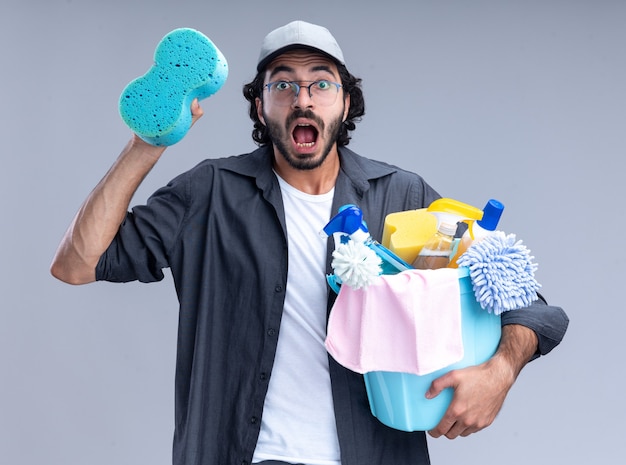 Hombre de limpieza guapo joven asustado con camiseta y gorra sosteniendo un cubo de herramientas de limpieza y una esponja aislada en la pared blanca