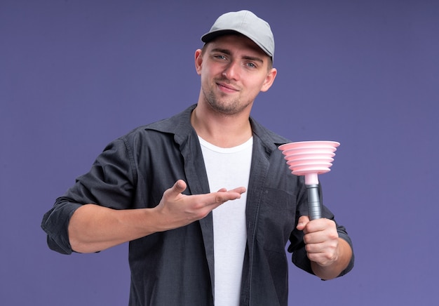 Hombre de limpieza guapo joven complacido con camiseta y gorra sosteniendo puntos ans en el émbolo aislado en la pared púrpura