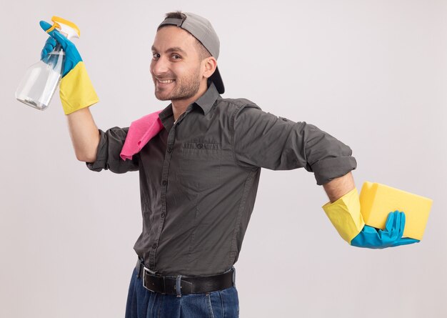 Hombre de limpieza joven alegre con ropa casual y gorra en guantes de goma sosteniendo spray de limpieza y una esponja con un trapo en su hombro feliz y alegre de pie sobre la pared naranja